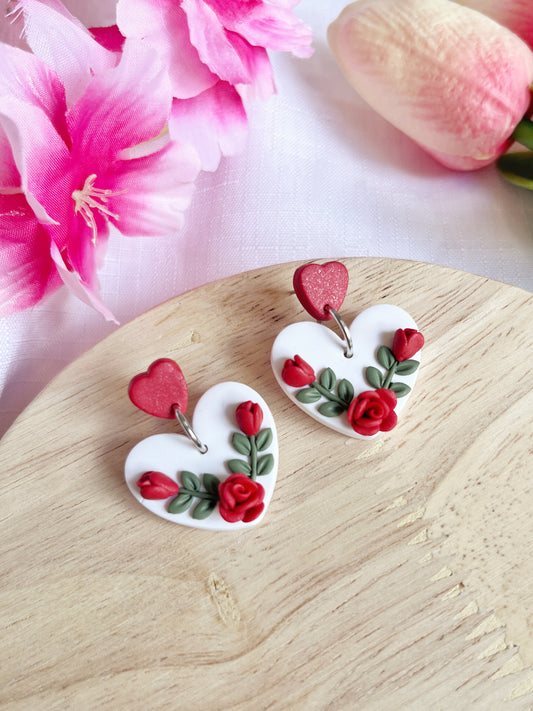 The Heart Rose Floral Polymer Clay Earrings by silverstar.nz showcase heart-shaped designs with red roses and green leaves on a wooden surface. Pink flowers and a tulip softly enhance the funky, floral aesthetic in the background.