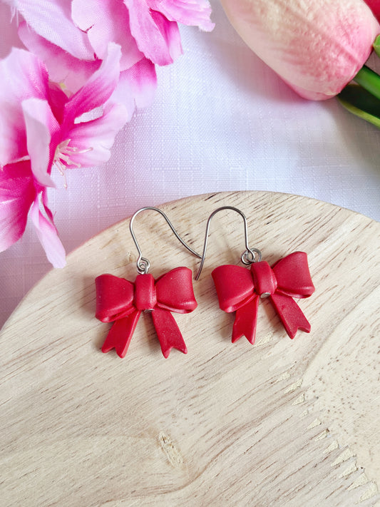 The Red Bow Polymer Clay Earrings by silverstar.nz, featuring stainless steel hooks, rest on a wooden surface. In the background, pink flowers and a tulip lie on white textured fabric, highlighting their handmade charm.