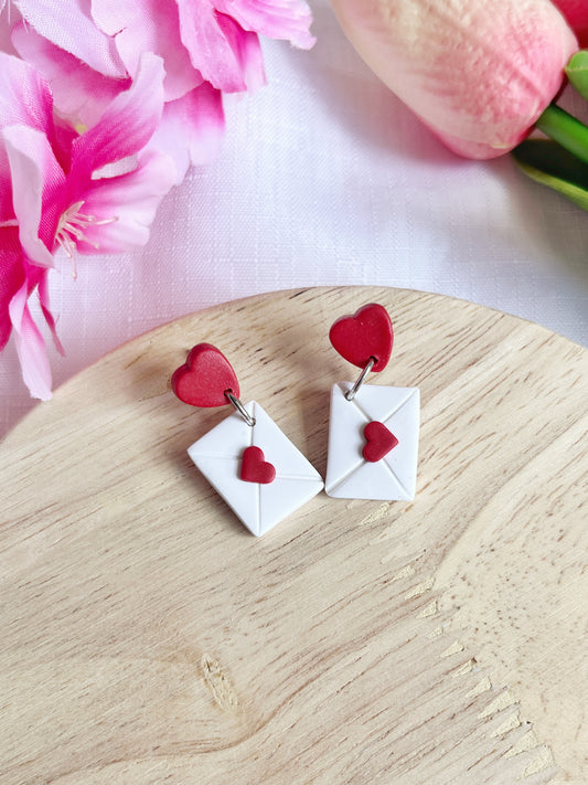 Valentines Envelope Polymer Clay Earrings by silverstar.nz are heart-shaped with red seals, displayed on a wooden surface. Enhanced with stainless steel hooks, theyre surrounded by pink flowers and a tulip on a white cloth background.