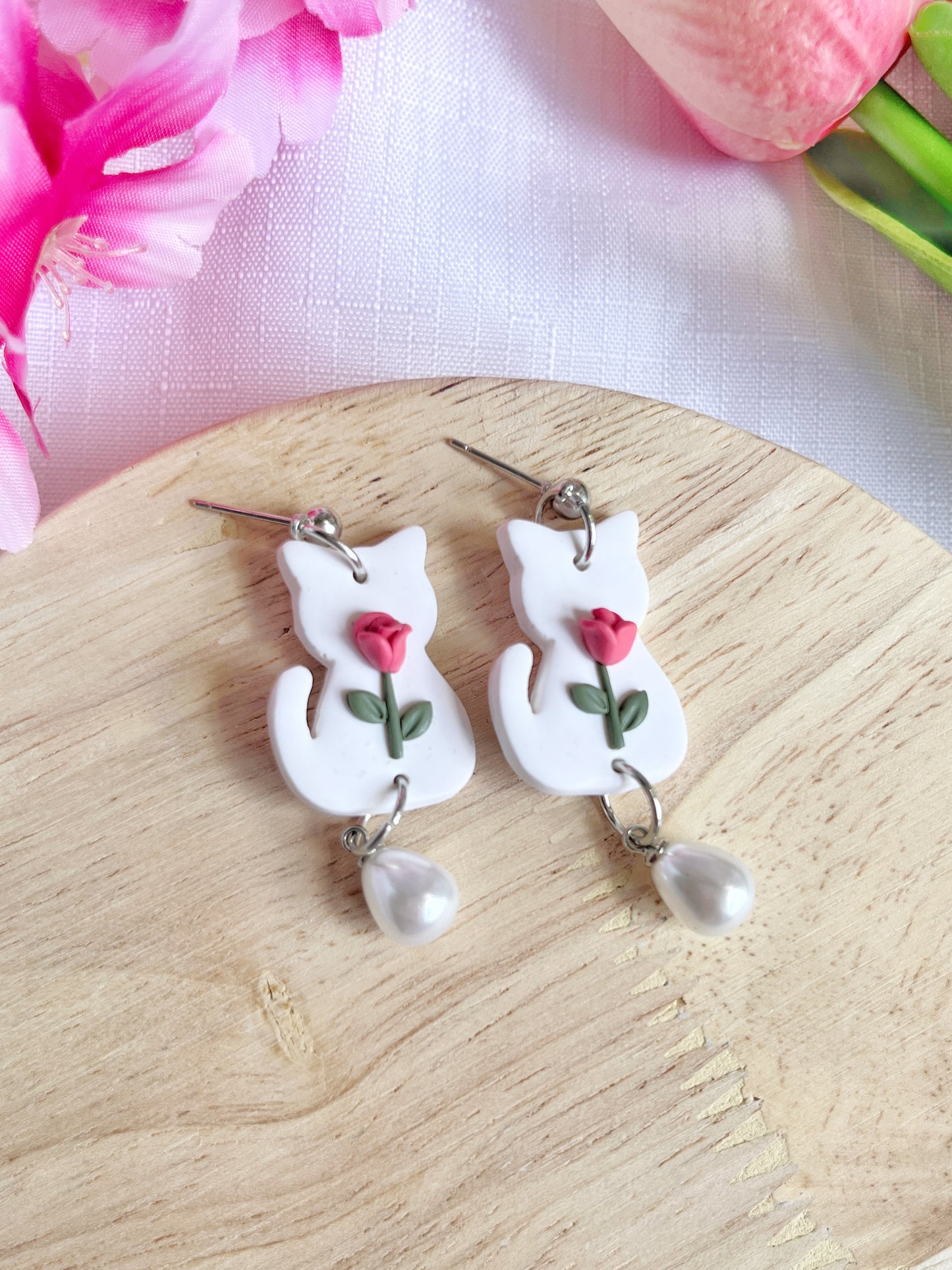 White Tulip Cat Earrings by silverstar.nz are crafted from polymer clay, featuring white cat shapes adorned with pink roses and dangling pearls. These handmade earrings rest on a wooden surface with pink flowers around, finished elegantly with stainless steel hooks.