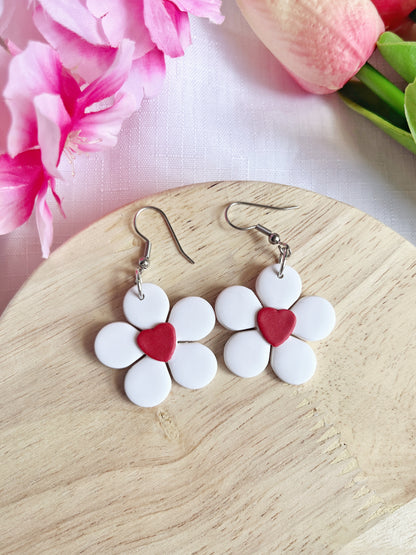 The Daisy Hearts Polymer Clay Earrings by silverstar.nz, featuring flower shapes with white petals and red heart centers, rest on a round wooden surface. Pink flowers and green leaves in the background enhance their charm.