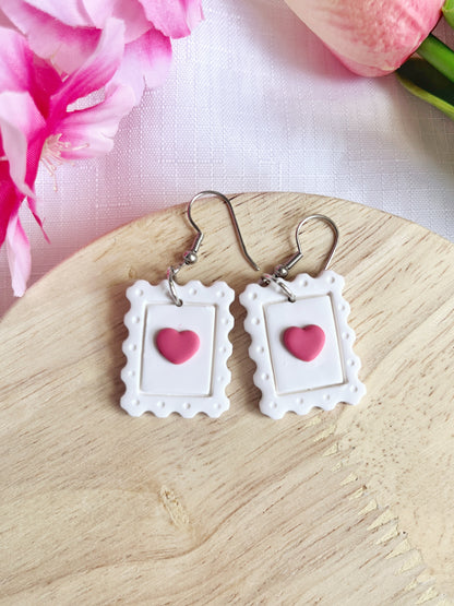 Heart Stamp Polymer Clay Earrings by silverstar.nz, shaped like white postage stamps with pink heart centers, dangle elegantly on stainless steel hooks on a wooden surface. Partial pink flowers in the background enhance the scenes charm.