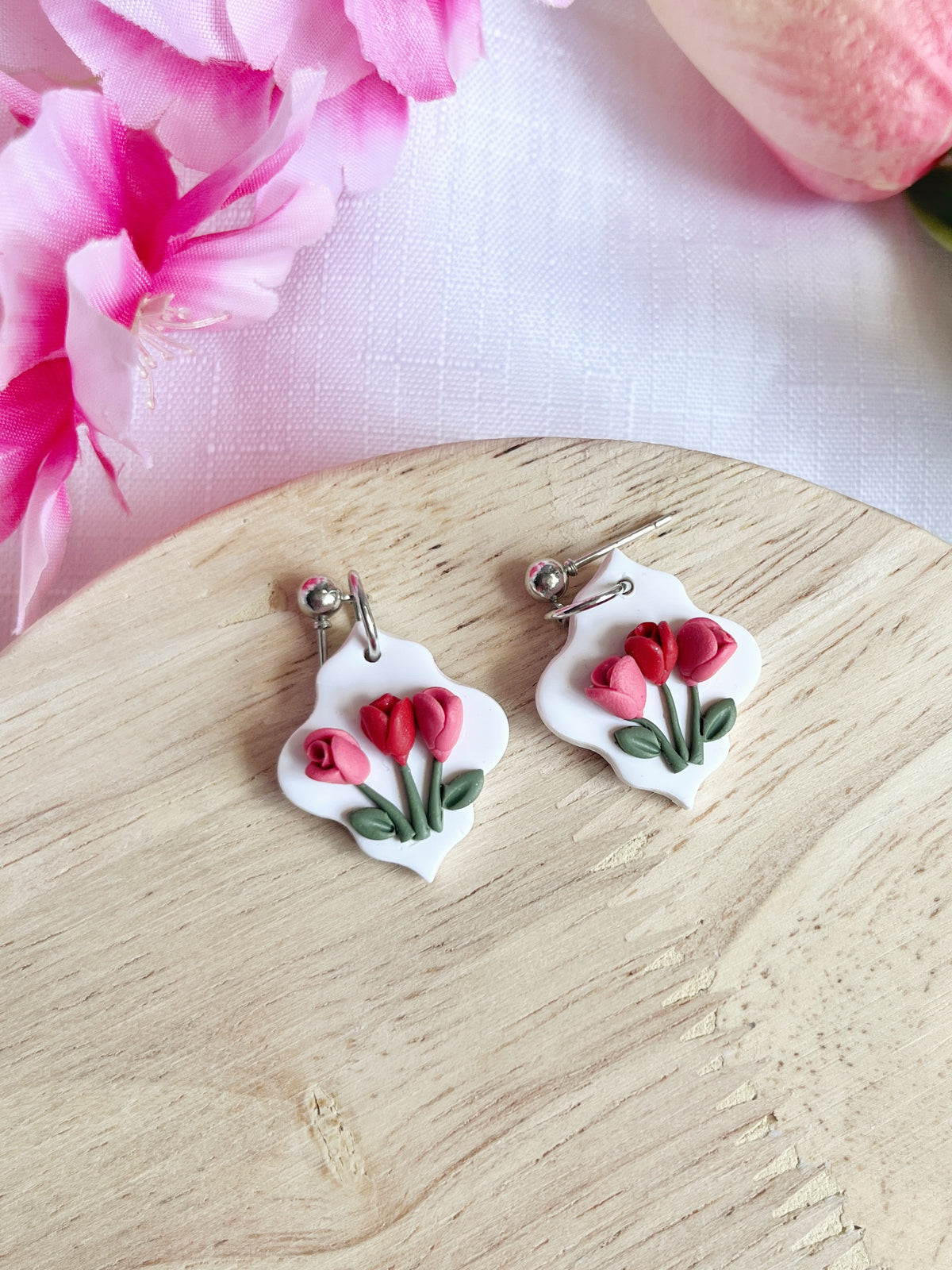 The Tulip Floral Polymer Clay Earrings by silverstar.nz, with delicate pink and red flowers, rest gracefully on a wooden surface. Their stainless-steel hooks guarantee durability, complemented by nearby pink flowers adding a soft, floral touch to the scene.