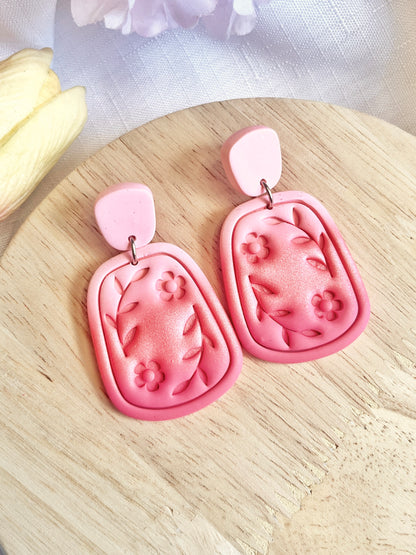 Statement Floral Polymer Clay Earrings from silverstar.nz feature a unique design with pink floral and leaf patterns on a rectangular shape with rounded edges, set against a wooden surface. These handmade earrings showcase darker pink flower and leaf designs paired with a lighter pink top piece.