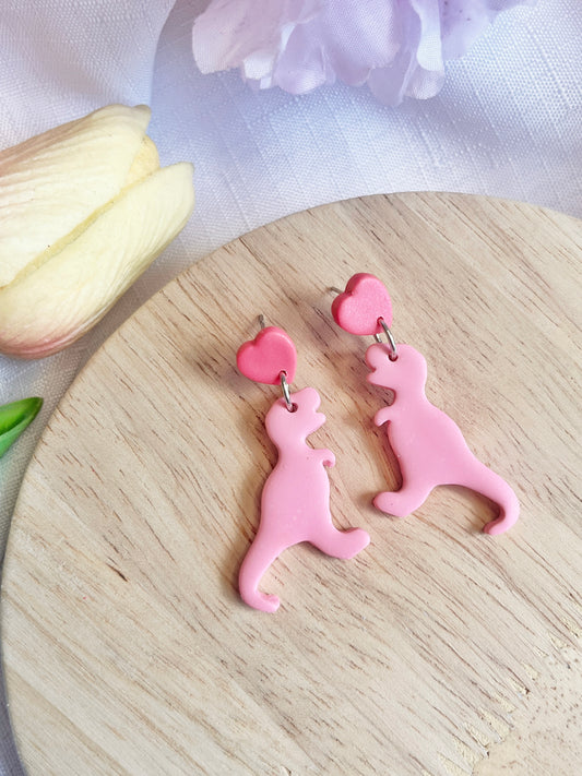 The Heart Dino Polymer Clay Earrings by silverstar.nz, shaped like pink dinosaurs with heart studs, rest on a wooden surface. A softly-focused white and pink flower in the background highlights their unique design.