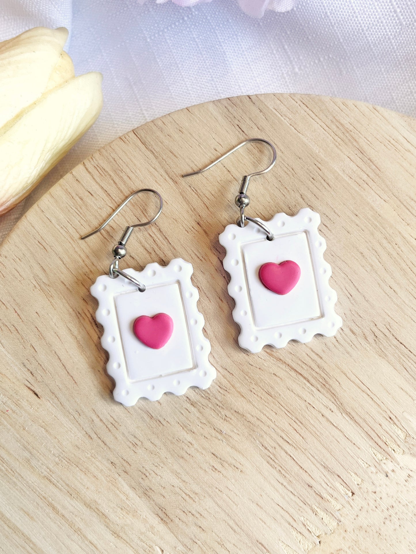 The Heart Stamp Polymer Clay Earrings by silverstar.nz, featuring pink hearts in white postage stamp frames, rest elegantly on a light wooden surface and are crafted with stainless steel hooks for lasting wear.