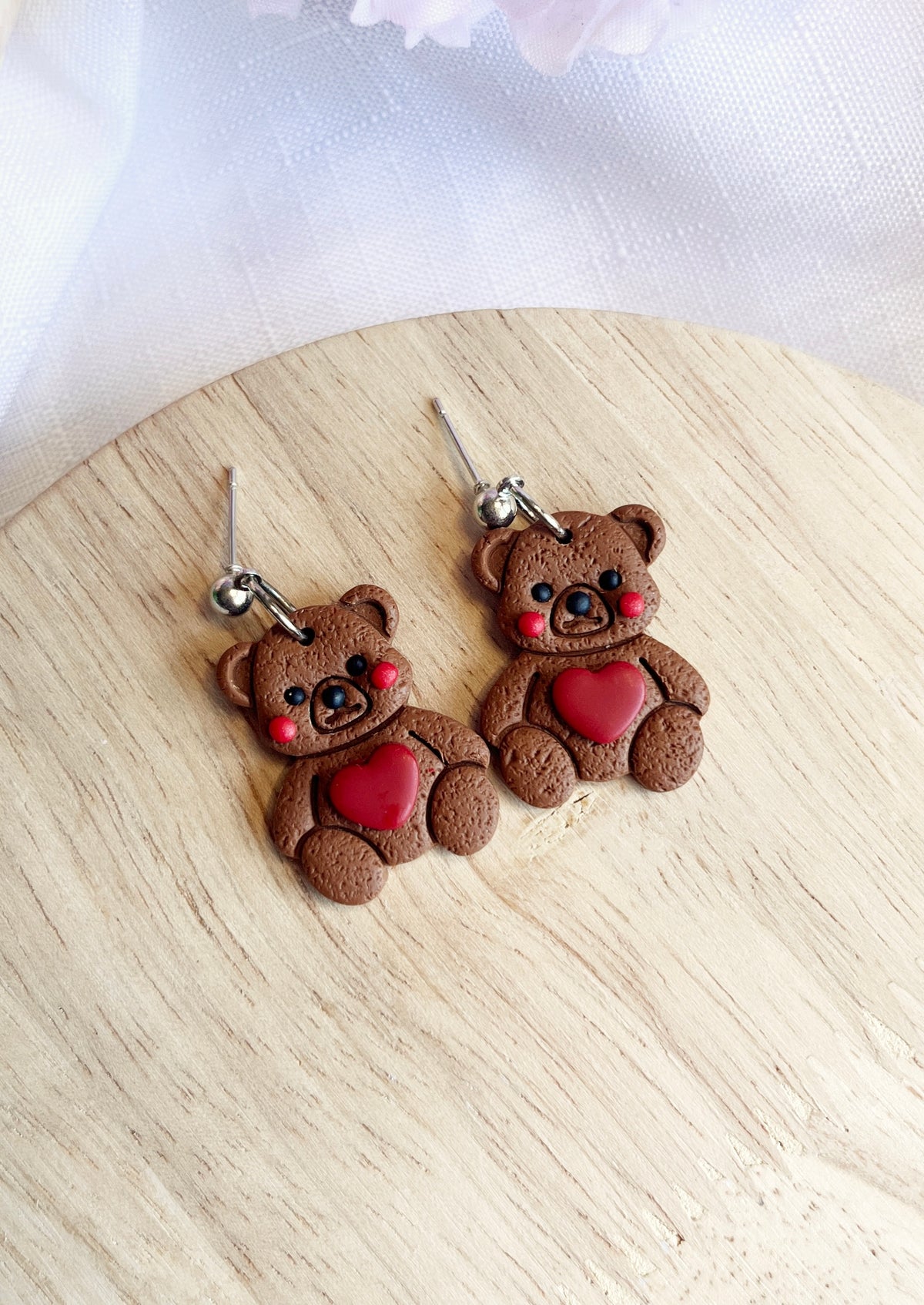 The Love Teddy Bear Polymer Clay Earrings by silverstar.nz, featuring smiling bears with rosy cheeks and red heart details, are displayed on a wooden surface. Crafted from polymer clay, each earring is adorned with stainless steel hooks for an adorable handmade accessory.
