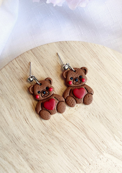 The Love Teddy Bear Polymer Clay Earrings by silverstar.nz, featuring smiling bears with rosy cheeks and red heart details, are displayed on a wooden surface. Crafted from polymer clay, each earring is adorned with stainless steel hooks for an adorable handmade accessory.