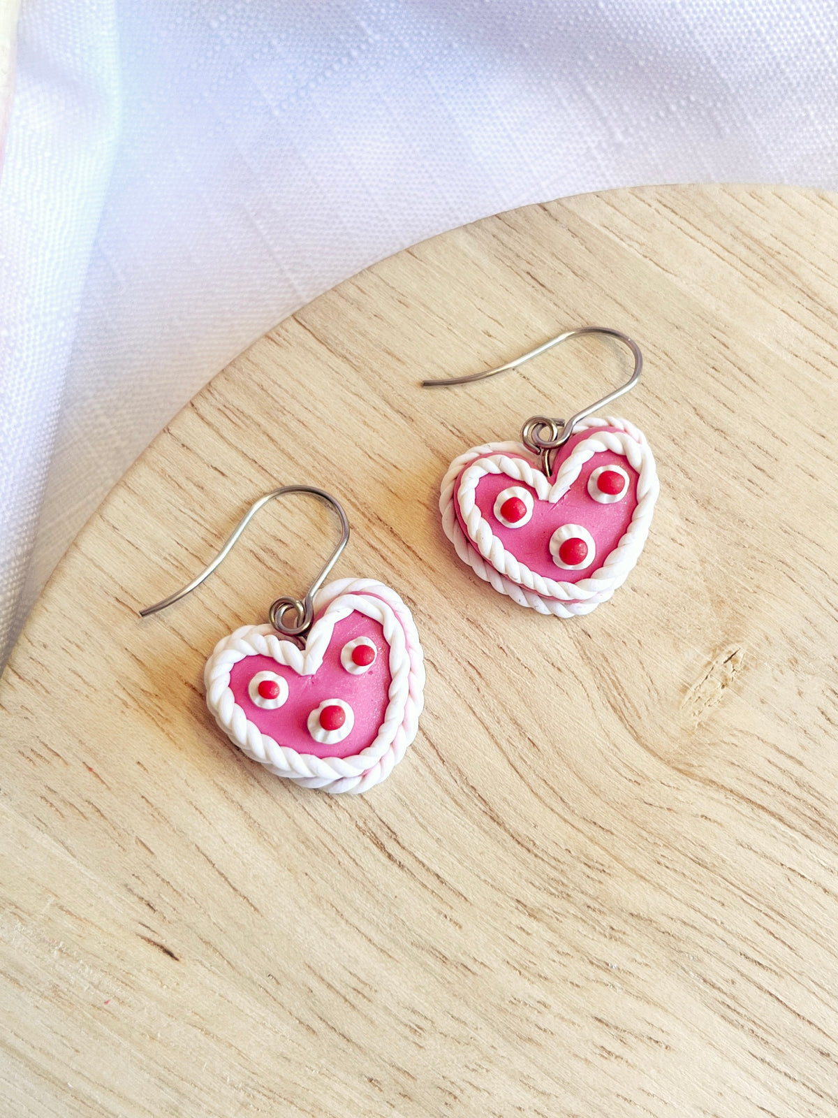 The silverstar.nz Vintage Heart Cake Polymer Clay Earrings feature pink centers, white rope edges, and tiny white flowers. Crafted from polymer clay with stainless steel hooks, these handmade heart-shaped earrings are elegantly displayed on a wooden surface, showcasing artisanal skill.