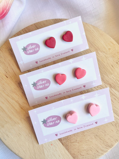 Displayed on a round wooden board, three pairs of valentines heart polymer clay stud earrings by silverstar.nz feature funky red and pink designs. The cards state handmade in New Zealand and include durable stainless steel hooks.
