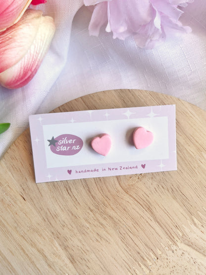 The Valentines Heart Polymer Clay Stud Earrings by silverstar.nz feature a funky design in light pink and hang from stainless steel hooks, displayed on a silver star.nz handmade in New Zealand card resting on a wooden surface with nearby pink and white flowers.