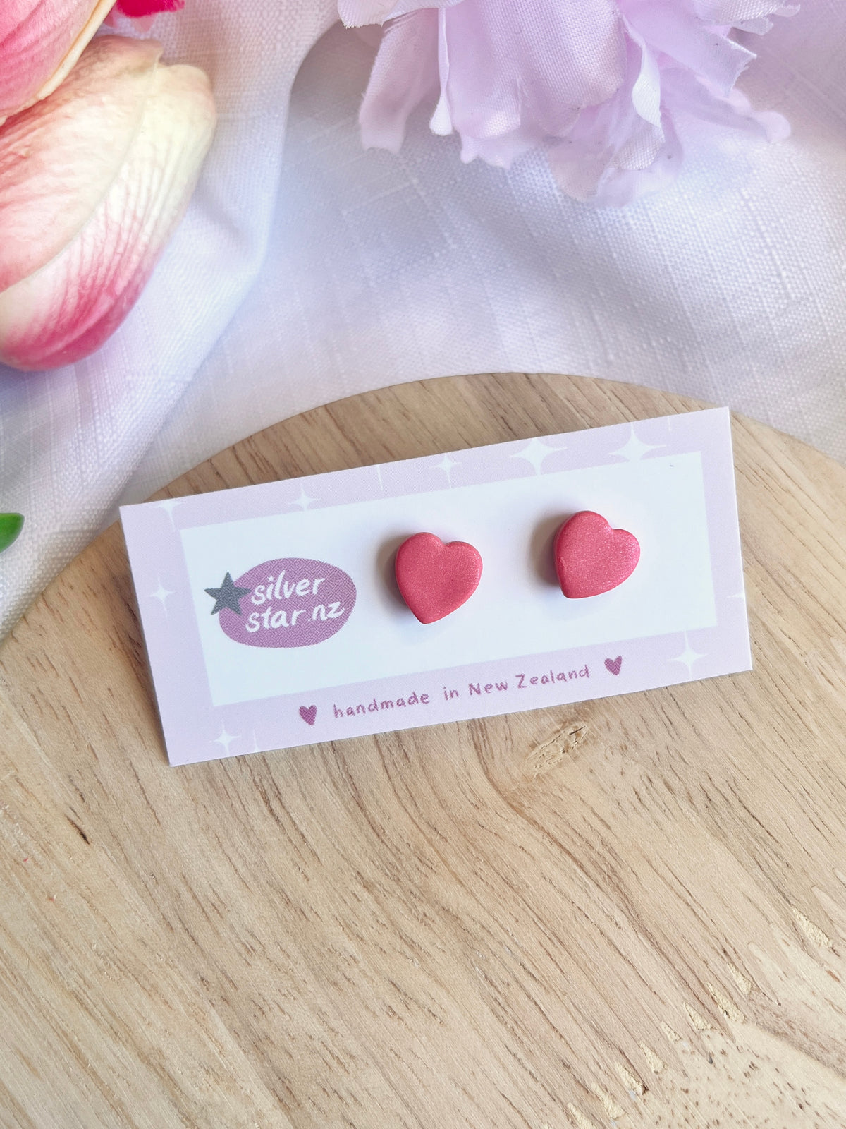 A pair of Valentines Heart Polymer Clay Stud Earrings from silverstar.nz, showcasing their New Zealand origin, rests on a card. These funky earrings display pastel flowers on handmade clay with stainless steel hooks, set against a wooden surface.