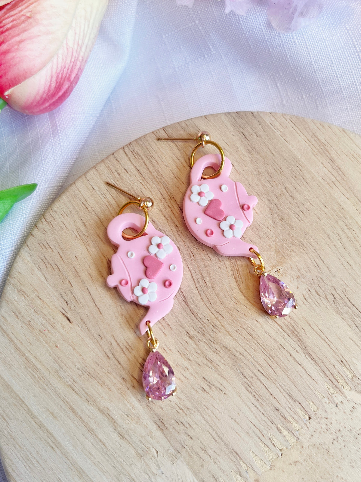 The Love Tea Pot Polymer Clay Earrings from silverstar.nz are pink tea pot-shaped with white flower details and dangling pink teardrop gems, elegantly displayed on a wooden surface. A soft fabric and pink flower in the background accentuate their unique handmade design.