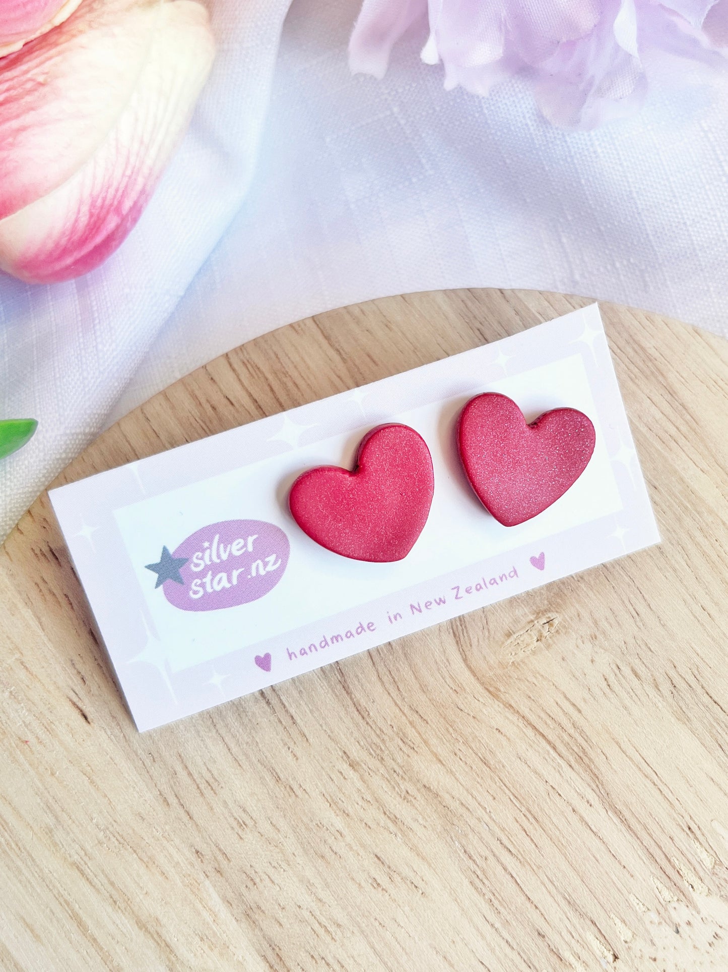 The Valentines Heart Polymer Clay Stud Earrings by silverstar.nz, featuring pink heart shapes and stainless steel hooks, are displayed on a handmade in New Zealand card. They rest elegantly on light wood with soft fabric and flower petals in the background.