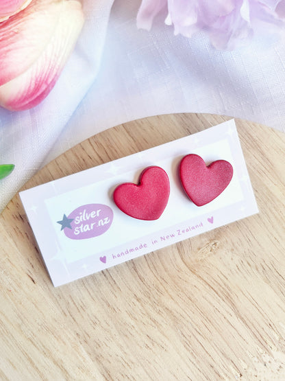 The Valentines Heart Polymer Clay Stud Earrings by silverstar.nz, featuring pink heart shapes and stainless steel hooks, are displayed on a handmade in New Zealand card. They rest elegantly on light wood with soft fabric and flower petals in the background.