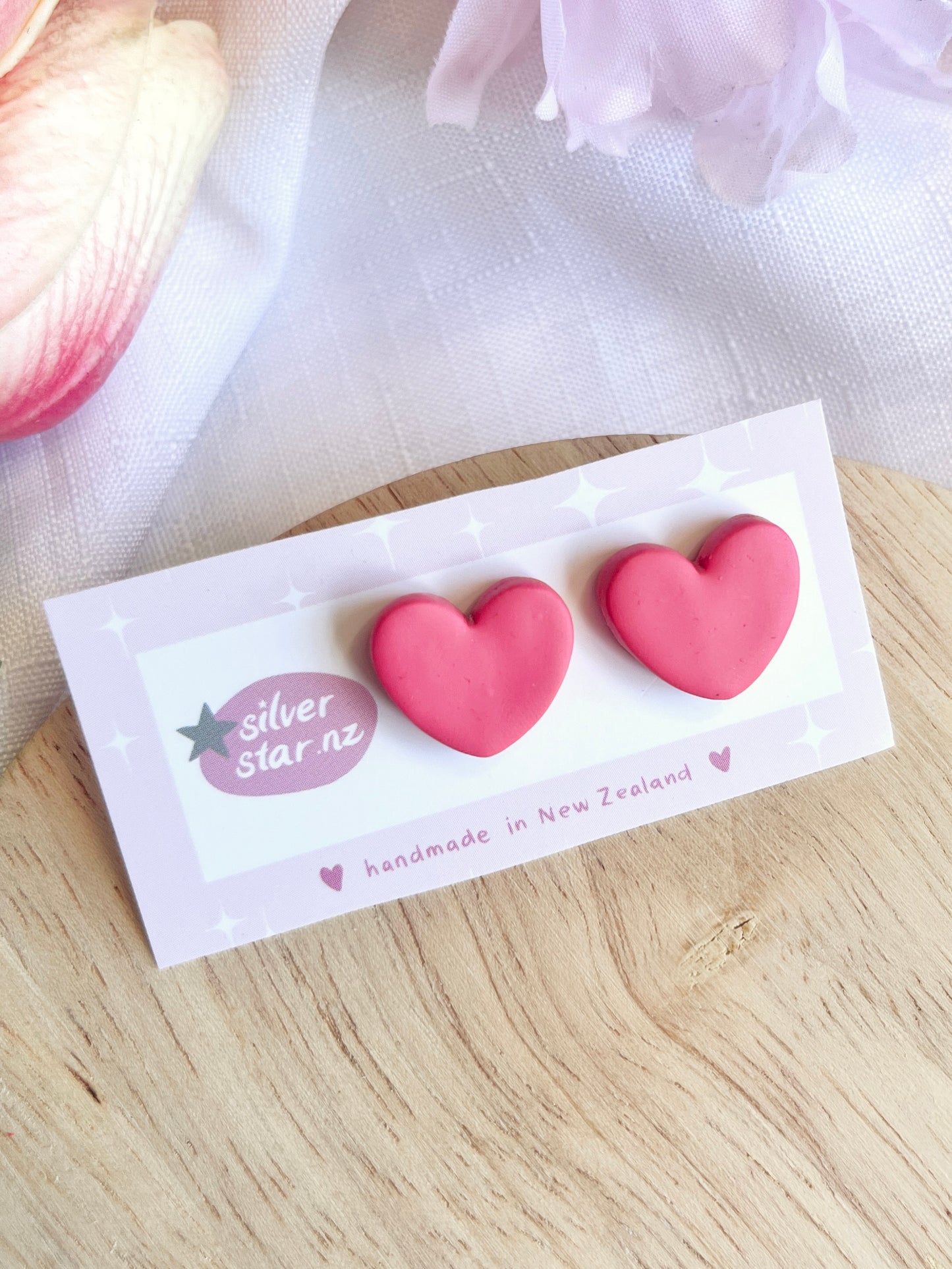 On a wooden surface, a card presents two pink heart-shaped earrings with stainless steel hooks and funky designs. The card reads silverstar.nz and handmade in New Zealand, adorned with star illustrations. A soft pink flower is partially visible nearby.