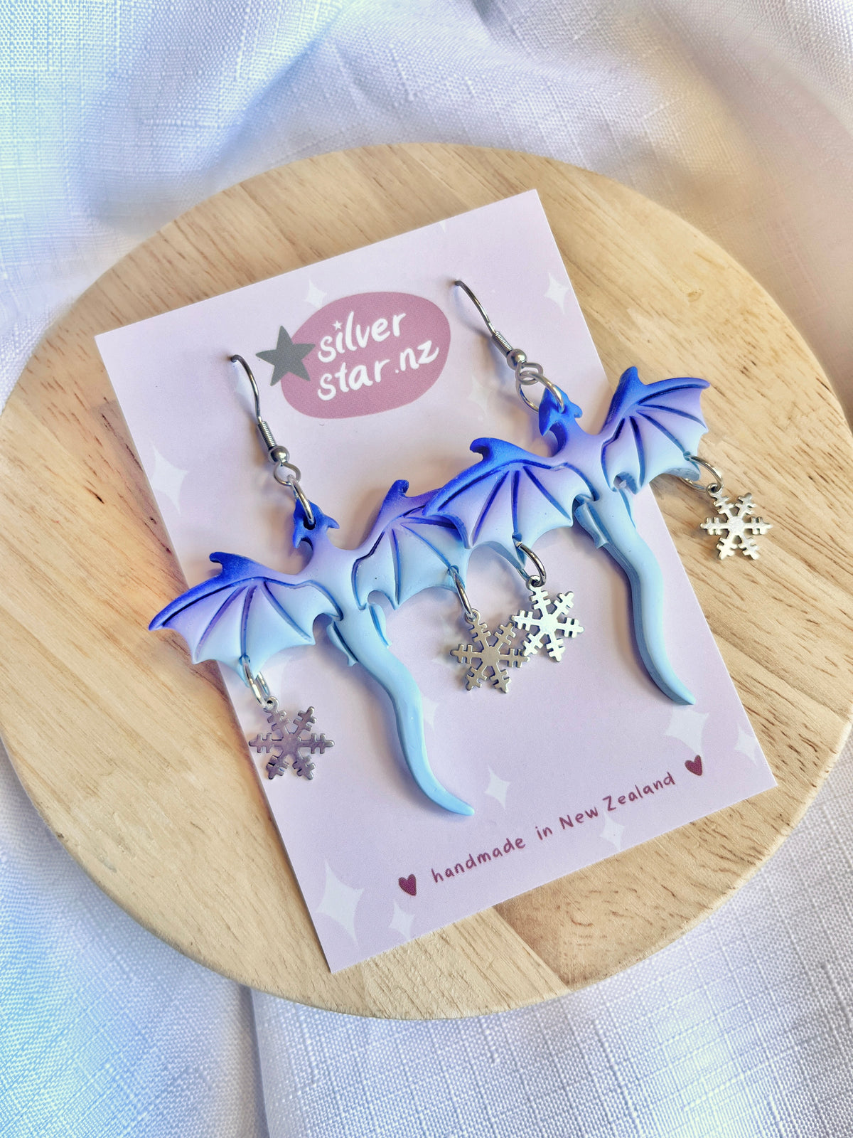 The Winter Snow Dragon Polymer Clay Earrings by silverstar.nz feature intricate blue dragon designs and dangling silver snowflakes, crafted with polymer clay and set on stainless steel hooks. Displayed on wood, the card reads silver star.nz and handmade in New Zealand.
