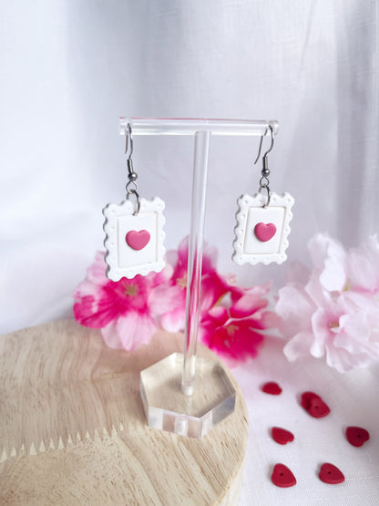Heart Stamp Polymer Clay Earrings by silverstar.nz, styled as white postage stamps with red heart centers, are showcased on a clear stand. The scene includes pink flowers on a light wooden surface with small red heart decorations nearby.