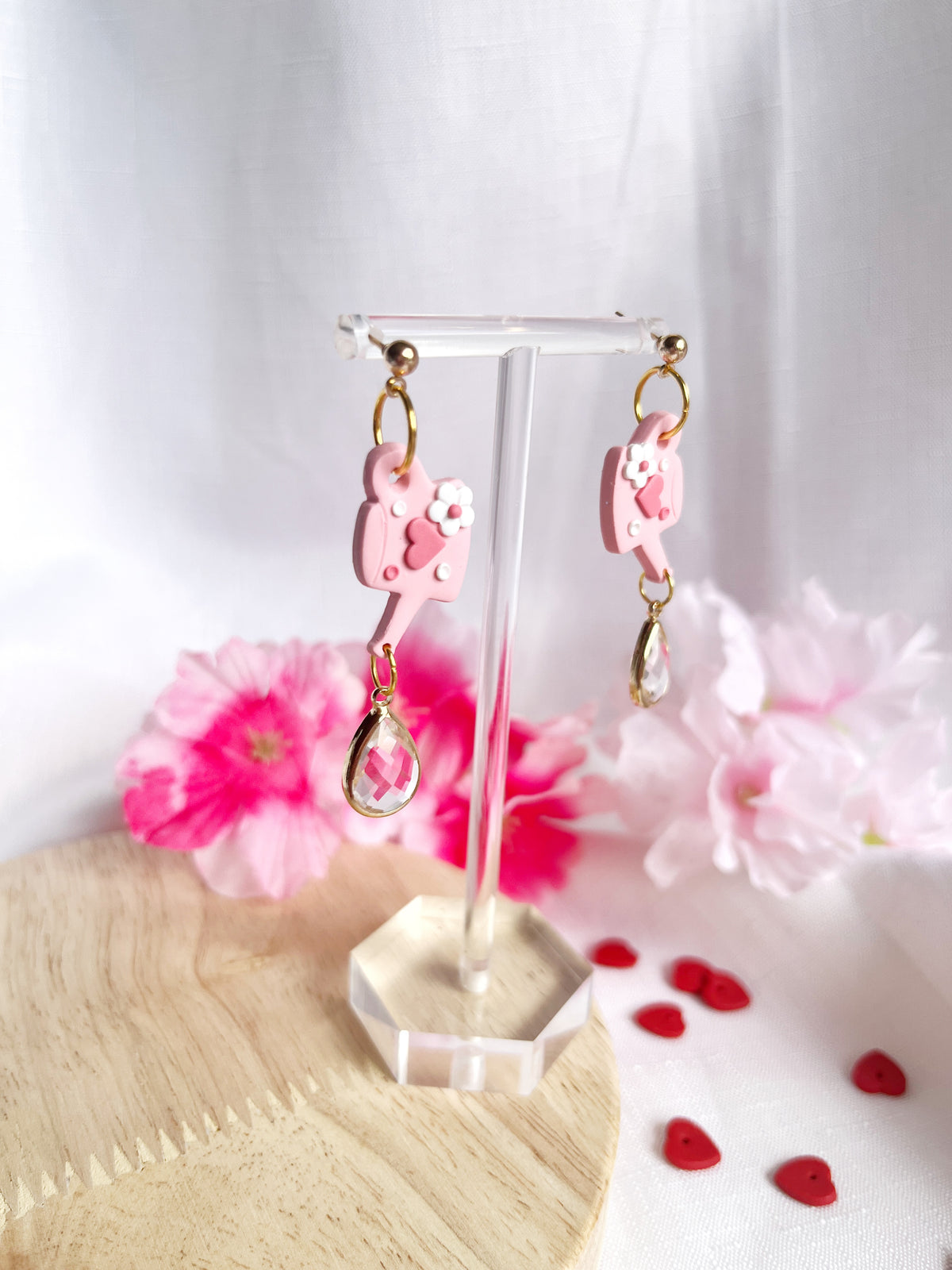 The Love Watering Can Polymer Clay Earrings by silverstar.nz elegantly dangle on a clear display stand, with a backdrop of pink flowers and red heart decorations on a wooden surface with white fabric.