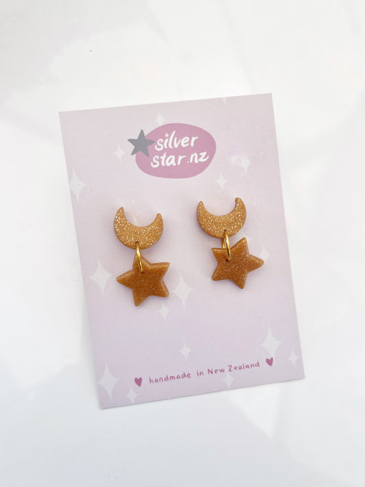 The Star + Moon Polymer Clay Earrings, with a glittery crescent moon and star design, hang from stainless steel hooks. Displayed on a card, they highlight the brand silverstar.nz and proudly note their handmade origin in New Zealand.