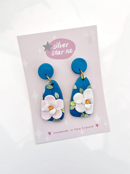 The Blue Floral Polymer Clay Earrings feature blue clay with white flowers, attached to a pale pink Silver Star card. The card displays silverstar.nz in a speech bubble and reads handmade in New Zealand. They include stainless steel hooks for enduring elegance.
