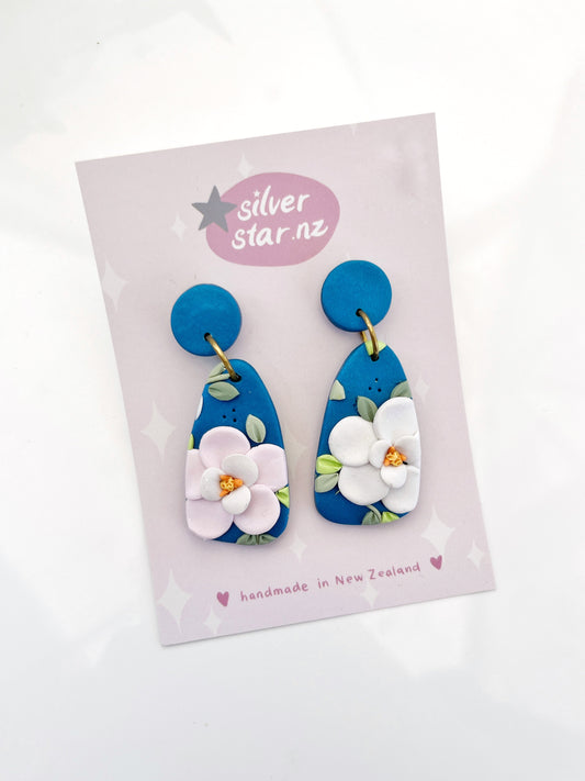 The Blue Floral Polymer Clay Earrings feature blue clay with white flowers, attached to a pale pink Silver Star card. The card displays silverstar.nz in a speech bubble and reads handmade in New Zealand. They include stainless steel hooks for enduring elegance.