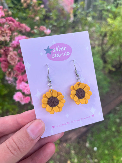 A hand showcases sunflower polymer clay earrings on a silverstar.nz branded card. The background features blurred pink flowers and greenery and the card states handmade in New Zealand.