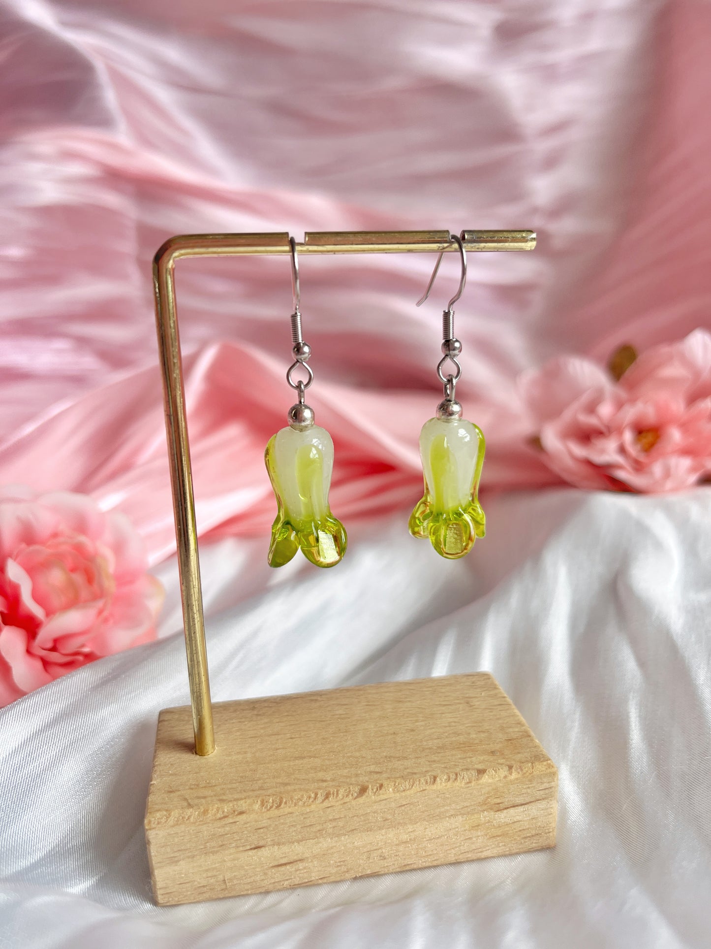 The Bok Choy Glass Bead Earrings by silverstar.nz, handmade and shaped like inverted flowers in green translucent glass, hang elegantly on a wooden stand. Theyre set against soft pink satin fabric with pink flowers, enhancing the delicate and feminine aesthetic.