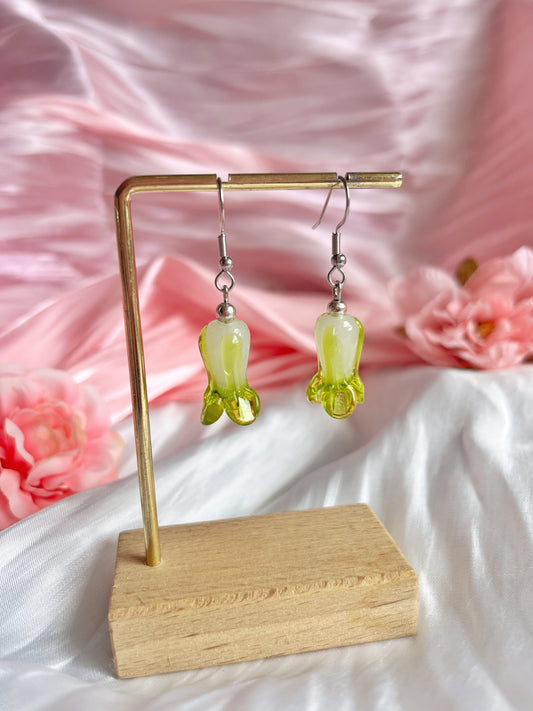 The Bok Choy Glass Bead Earrings by silverstar.nz, handmade and shaped like inverted flowers in green translucent glass, hang elegantly on a wooden stand. Theyre set against soft pink satin fabric with pink flowers, enhancing the delicate and feminine aesthetic.