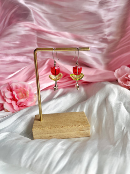 Displayed on a wooden stand, the Tulip Bead Earrings by silverstar.nz showcase unique red and yellow crescent shapes with dangling star charms and stainless steel hooks against a soft pink and white fabric backdrop adorned with pink rose accents.