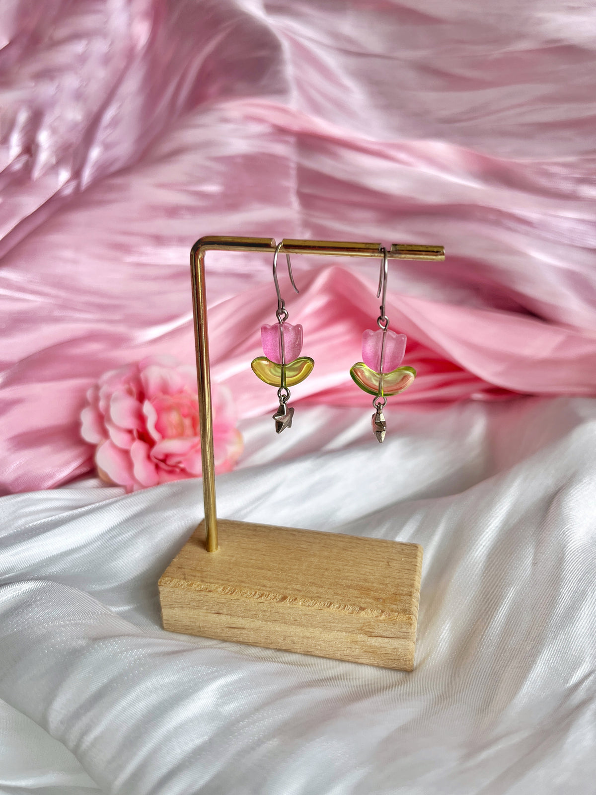 Tulip Bead Earrings by silverstar.nz: Pink and green handmade with dangling leaf charms on a wooden and metal stand. The earrings, set against soft pink and white satin fabric with a partially visible pink flower, feature unique craftsmanship with stainless steel hooks.