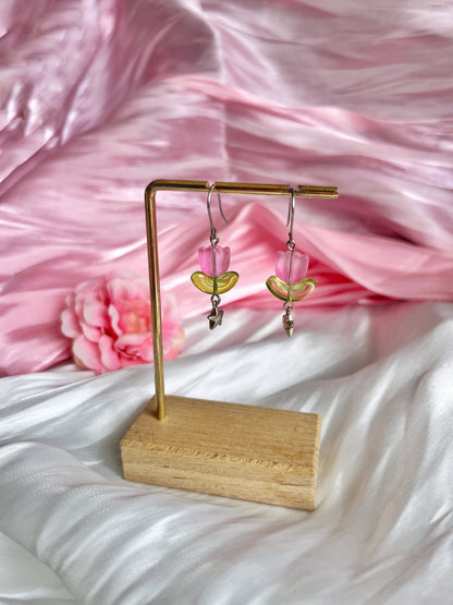Tulip Bead Earrings by silverstar.nz: Pink and green handmade with dangling leaf charms on a wooden and metal stand. The earrings, set against soft pink and white satin fabric with a partially visible pink flower, feature unique craftsmanship with stainless steel hooks.