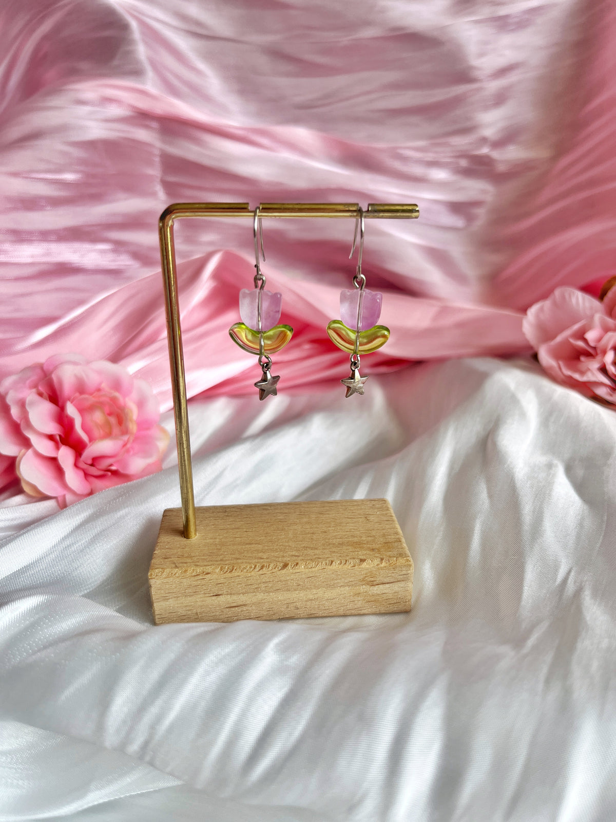 The Tulip Bead Earrings by silverstar.nz, featuring elegant crescent shapes and star pendants, hang on a gold stand against a backdrop of soft pink and white fabric adorned with pink flowers, creating a delicate and romantic atmosphere.