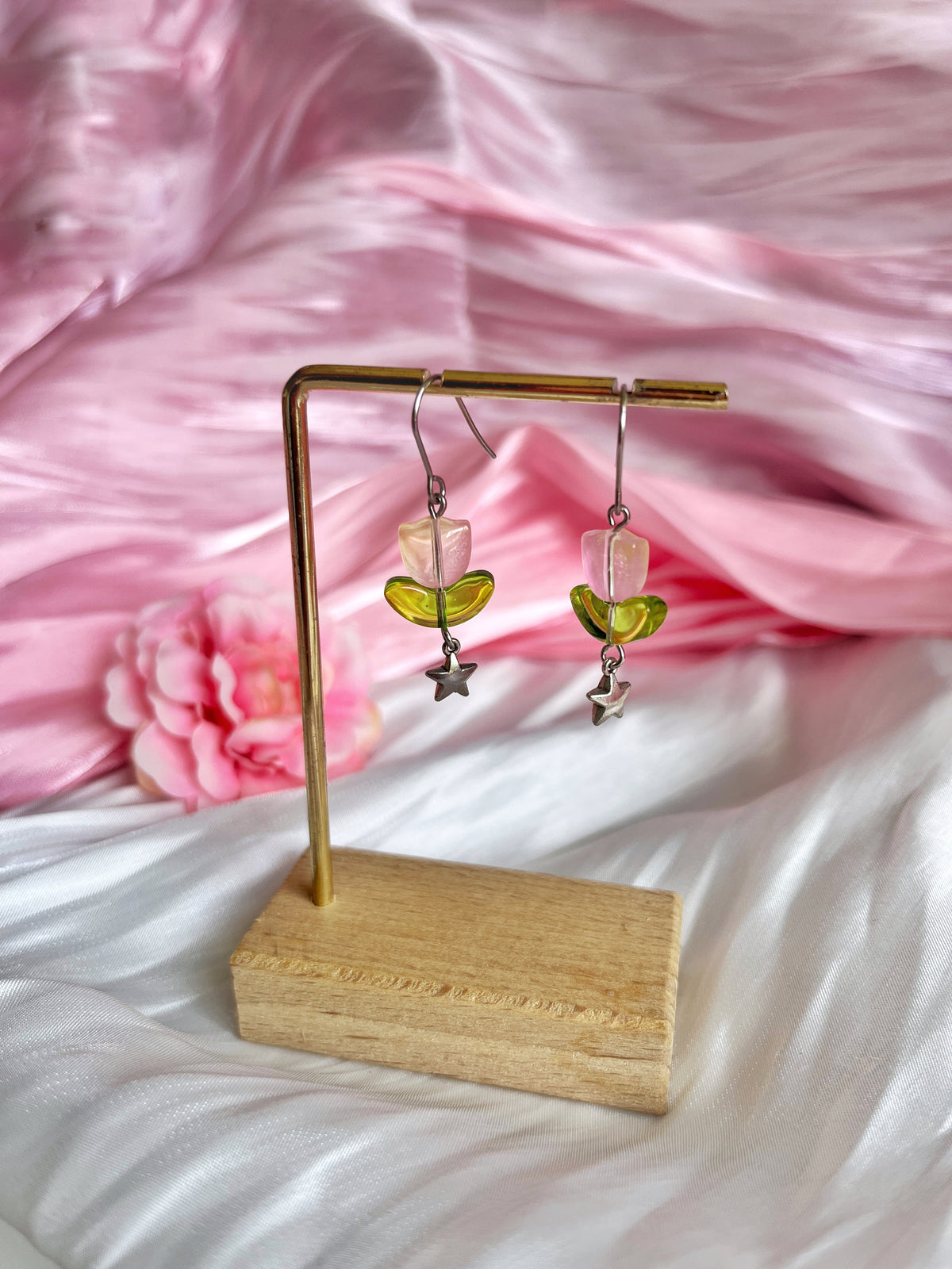 The Tulip Bead Earrings by silverstar.nz feature green half-circle shapes and star charms on stainless steel hooks. Displayed elegantly on a wooden stand, the earrings are set against a soft pink and white fabric backdrop with a pink flower decoration.