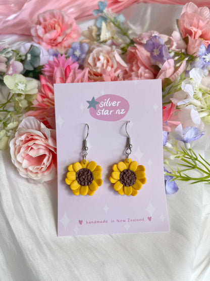Handmade Sunflower Polymer Clay Earrings by silverstar.nz with brown centers and yellow petals are displayed on a card. The backdrop features pink, purple, and white flowers.