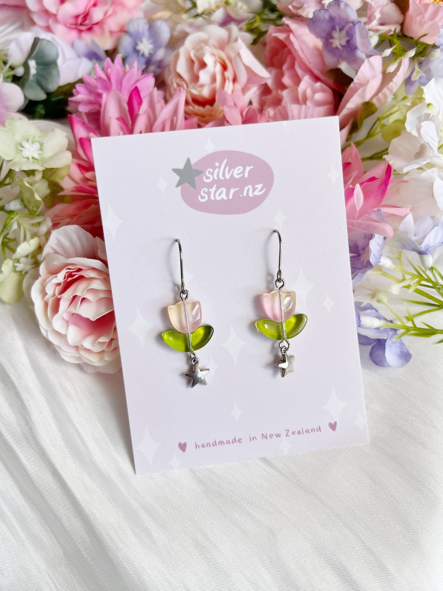 Tulip Bead Earrings have pink petals and green leaves, displayed on a silverstar.nz card. The handmade design uses stainless steel hooks and sits on a colorful flower backdrop, showcasing their unique appeal.