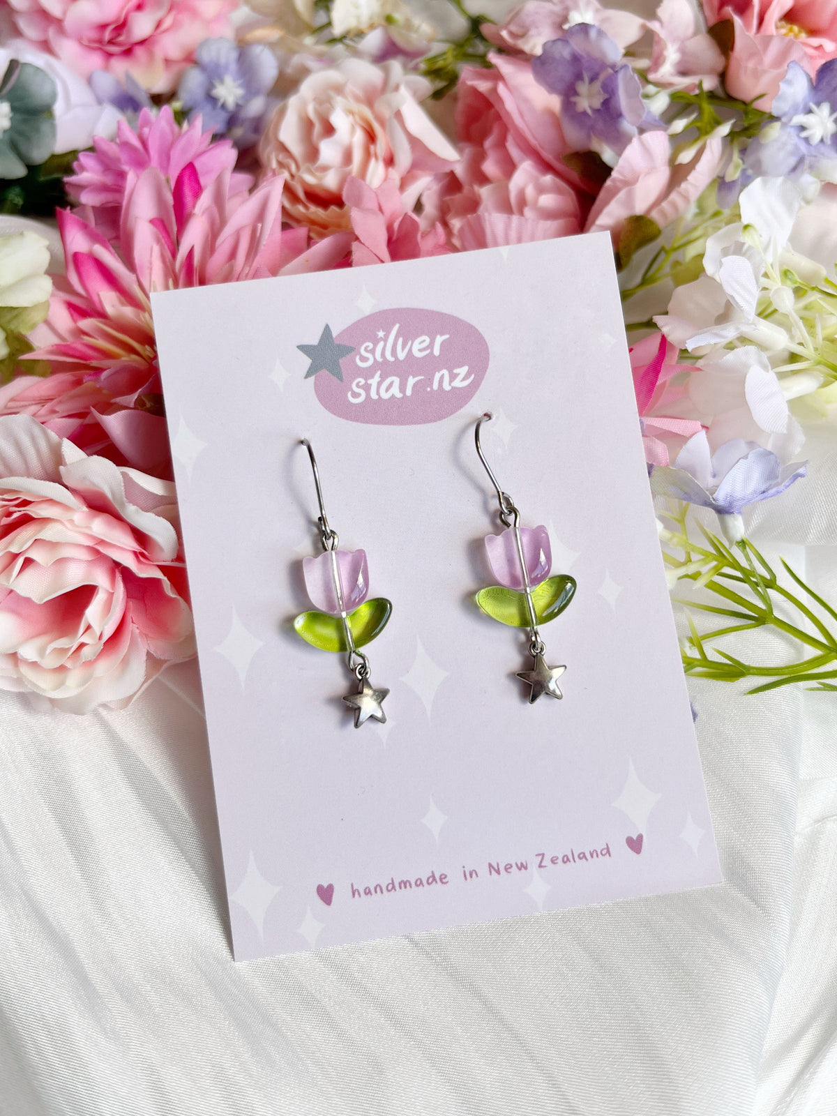 Tulip Bead Earrings by silverstar.nz showcase pink and green elements with silver stars on a soft-focus floral backdrop. Hanging from stainless steel hooks, they come displayed on a card labeled silver star nz.