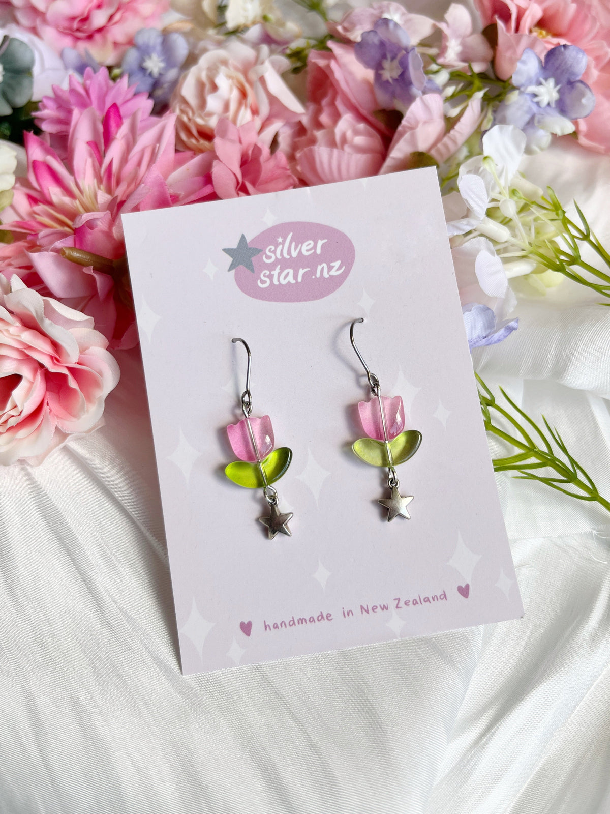 The Tulip Bead Earrings feature pink and green flowers with dangling silver stars, crafted with stainless steel hooks. They are displayed on a card reading silverstar.nz, handmade in New Zealand, against a background of pink, white, and purple flowers on a white surface.