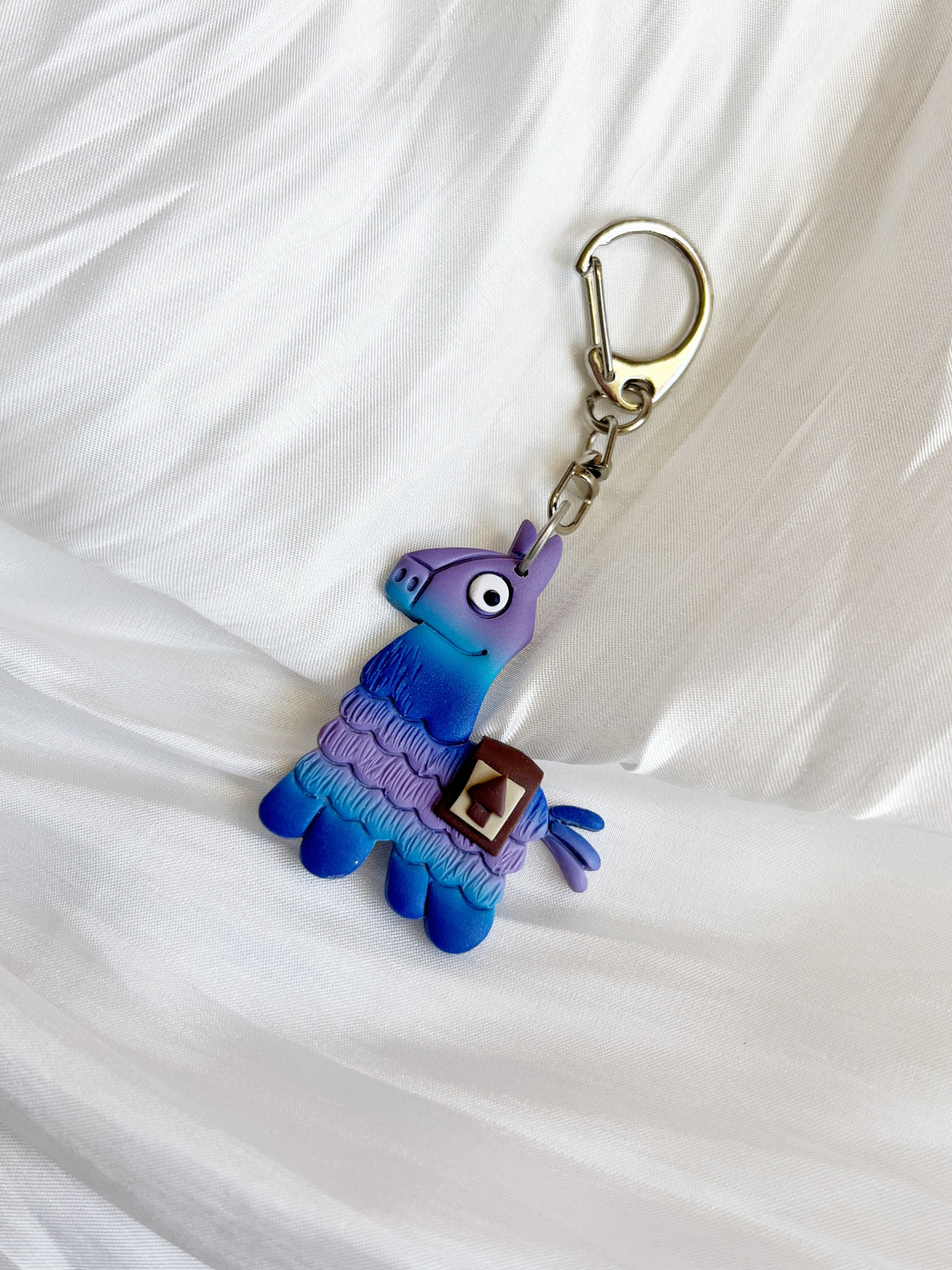 The Llama Polymer Clay Keyring by silverstar.nz features a vibrant blue and purple llama with a silver clasp, displayed on textured white fabric—an ideal unique gift choice.