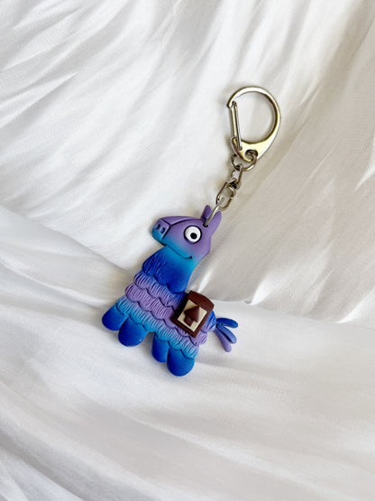 The Llama Polymer Clay Keyring by silverstar.nz features a vibrant blue and purple llama with a silver clasp, displayed on textured white fabric—an ideal unique gift choice.