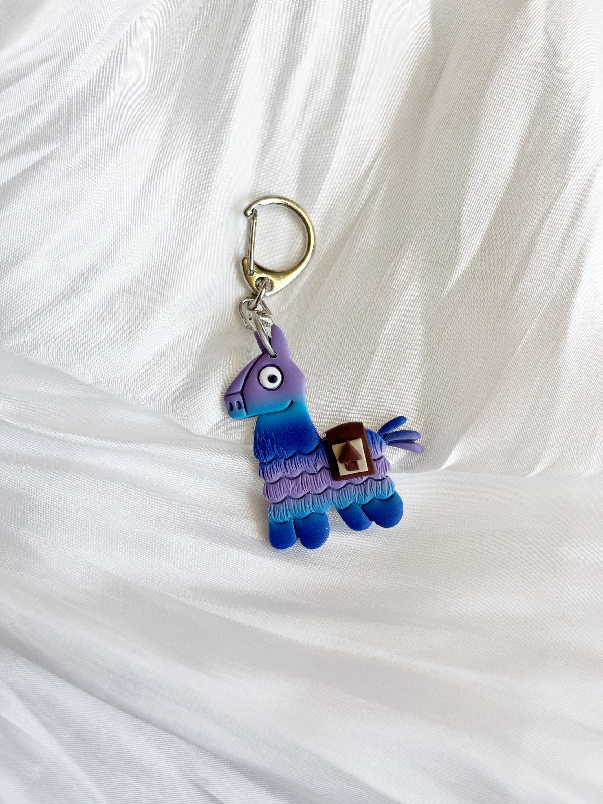The Llama Polymer Clay Keyring by silverstar.nz, shaped like a vibrant llama piñata in purple, blue, and teal, rests on white textured fabric—a charming pick for unique gifts.