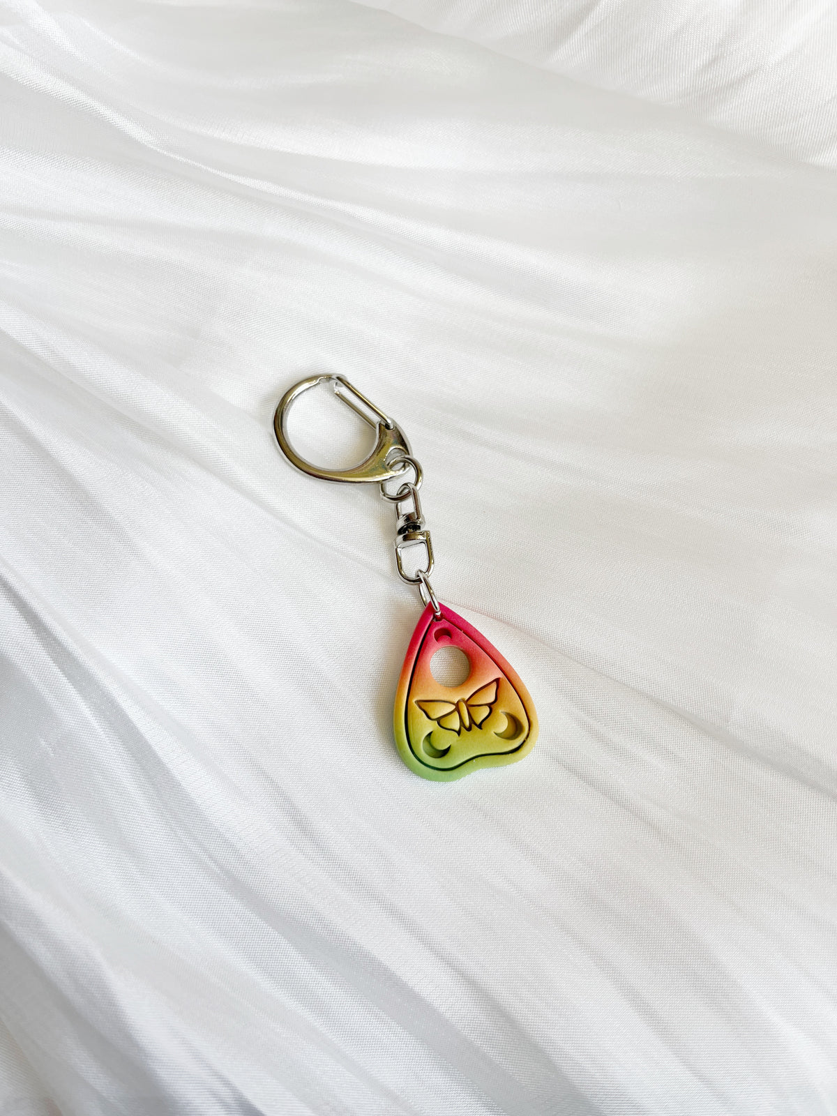 The Ouija Planchette Polymer Clay Keyring by silverstar.nz is a handmade New Zealand piece featuring a heart-shaped charm with a butterfly design and a gradient of red, yellow, and green on white textured fabric.