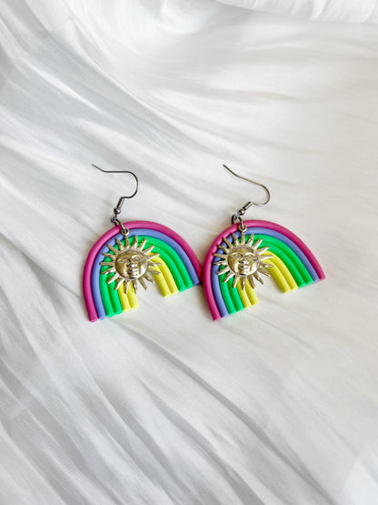 Silverstar.nzs Rainbow Arch Polymer Clay Earrings, featuring pink, blue, green, and yellow hues with silver sun charms, hang elegantly against a soft white fabric background for a unique design.