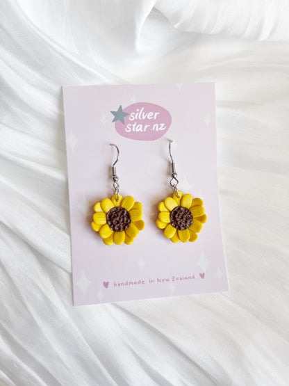 On a card labeled silverstar.nz, the Sunflower Polymer Clay Earrings, handmade in New Zealand with yellow petals and a brown center, are showcased against a soft white textured fabric.