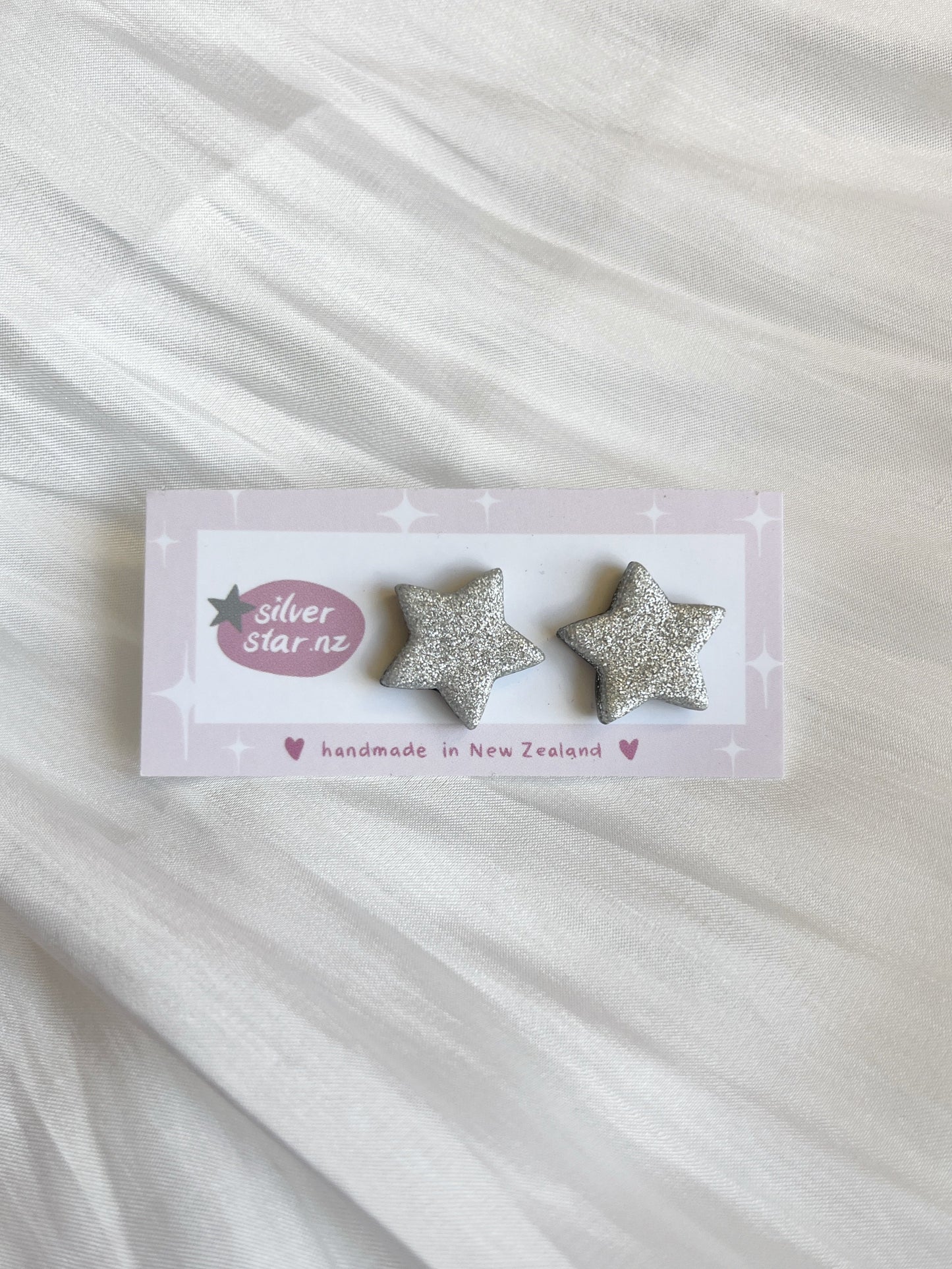 The Silver Star Polymer Clay Stud Earrings feature a unique glittery silver star design, displayed on a light pink card labeled silverstar.nz and handmade in New Zealand, resting elegantly on soft white fabric.