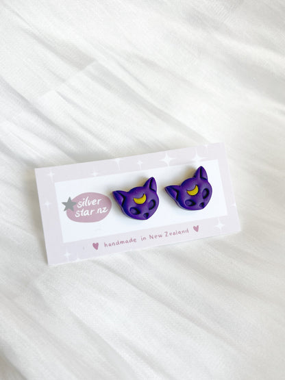 Purple Moon Cat Polymer Clay Stud Earrings by silverstar.nz, handmade in New Zealand, feature purple cat shapes with yellow crescent moons on their foreheads and are displayed on a branded card against a white fabric background.