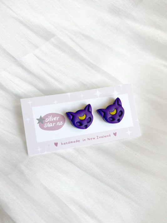Purple Moon Cat Polymer Clay Stud Earrings by silverstar.nz, handmade in New Zealand, feature purple cat shapes with yellow crescent moons on their foreheads and are displayed on a branded card against a white fabric background.