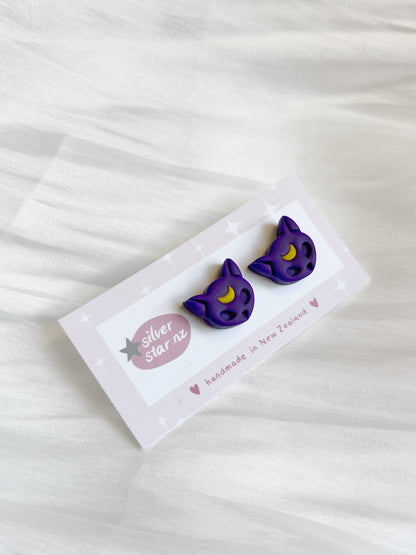Purple Moon Cat Polymer Clay Stud Earrings by silverstar.nz, featuring purple cat shapes with yellow crescent moons, displayed on a branded card against a white fabric background.