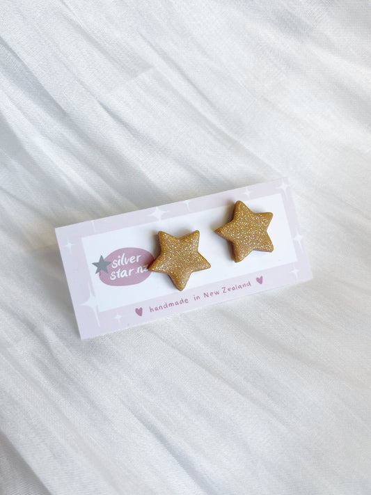 Two glittery gold star-shaped polymer clay earrings on a card labeled Gold Star, placed on white textured fabric. The card reads silverstar.nz and handmade in New Zealand with small decorative hearts, featuring stainless steel hooks.