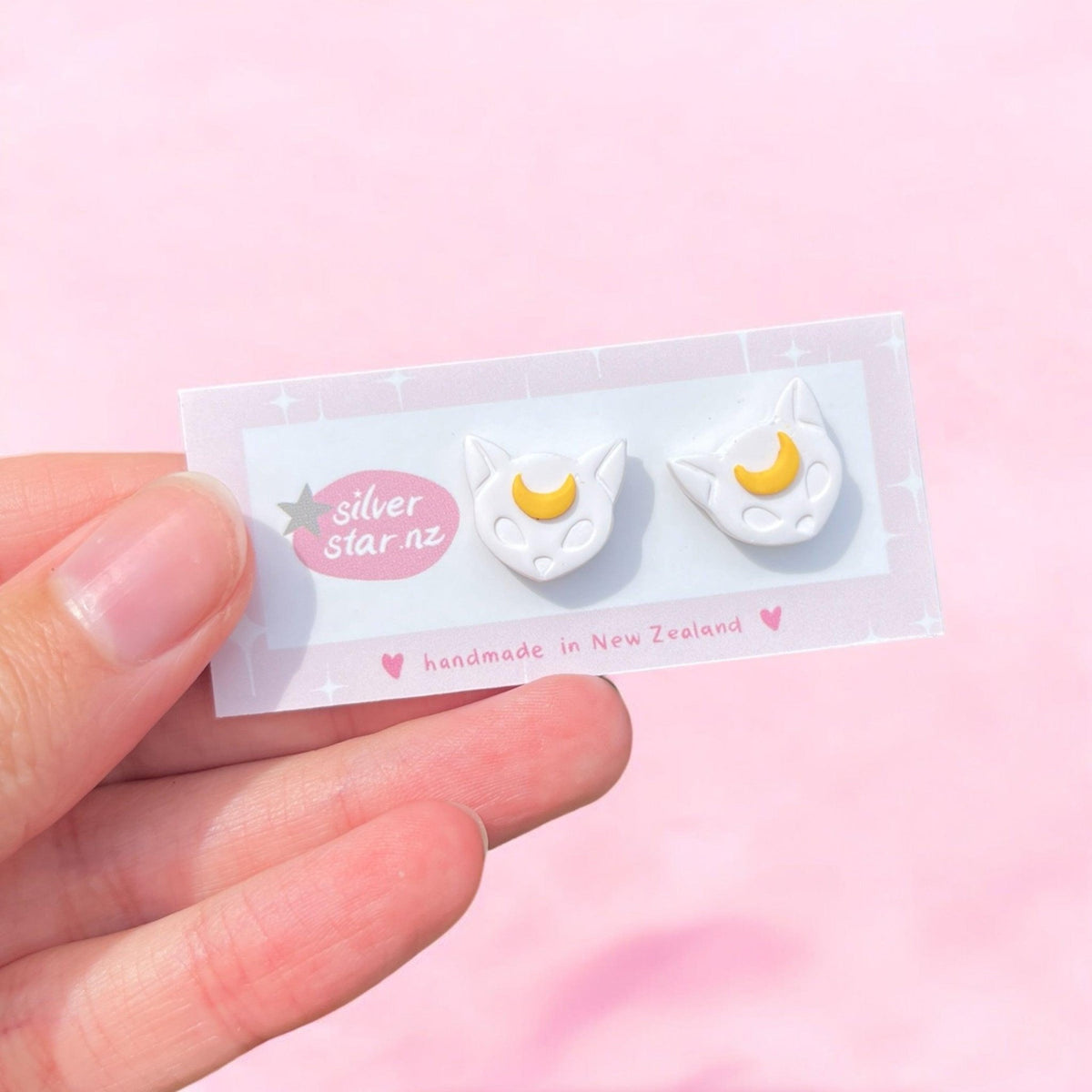 A hand holds a card showing Moon Cat Polymer Clay Stud Earrings—white cat-shaped studs with yellow crescent moons—from silverstar.nz, labeled "handmade in New Zealand," against a soft pink background.